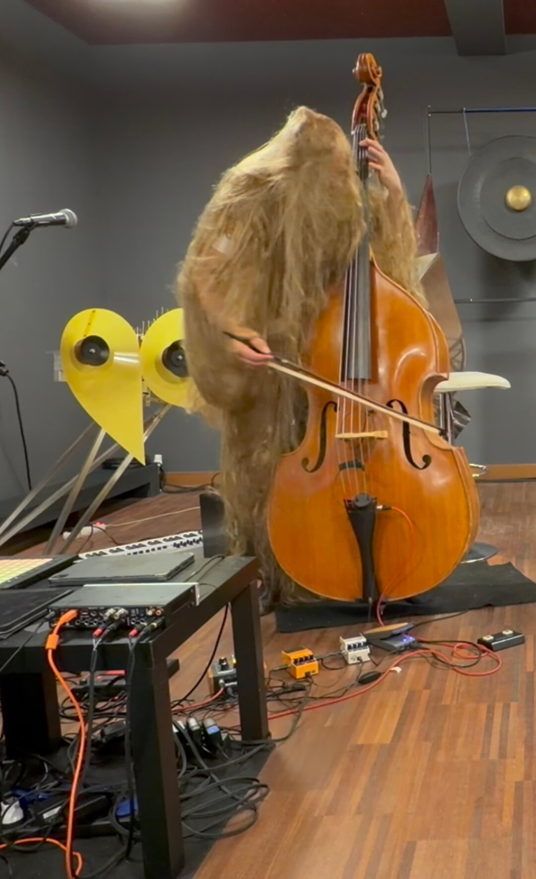 A performer in a shaggy costume plays the double bass; yellow objects and stage equipment are visible in the background.